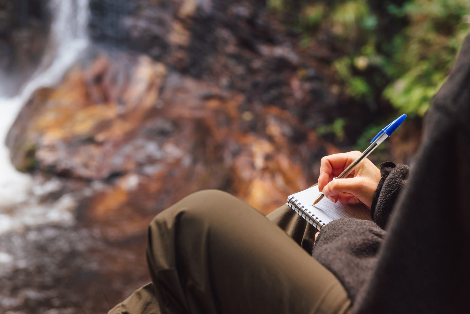 Person writing in a notebook by a stream, enjoying outdoor adventure and fishing lifestyle.